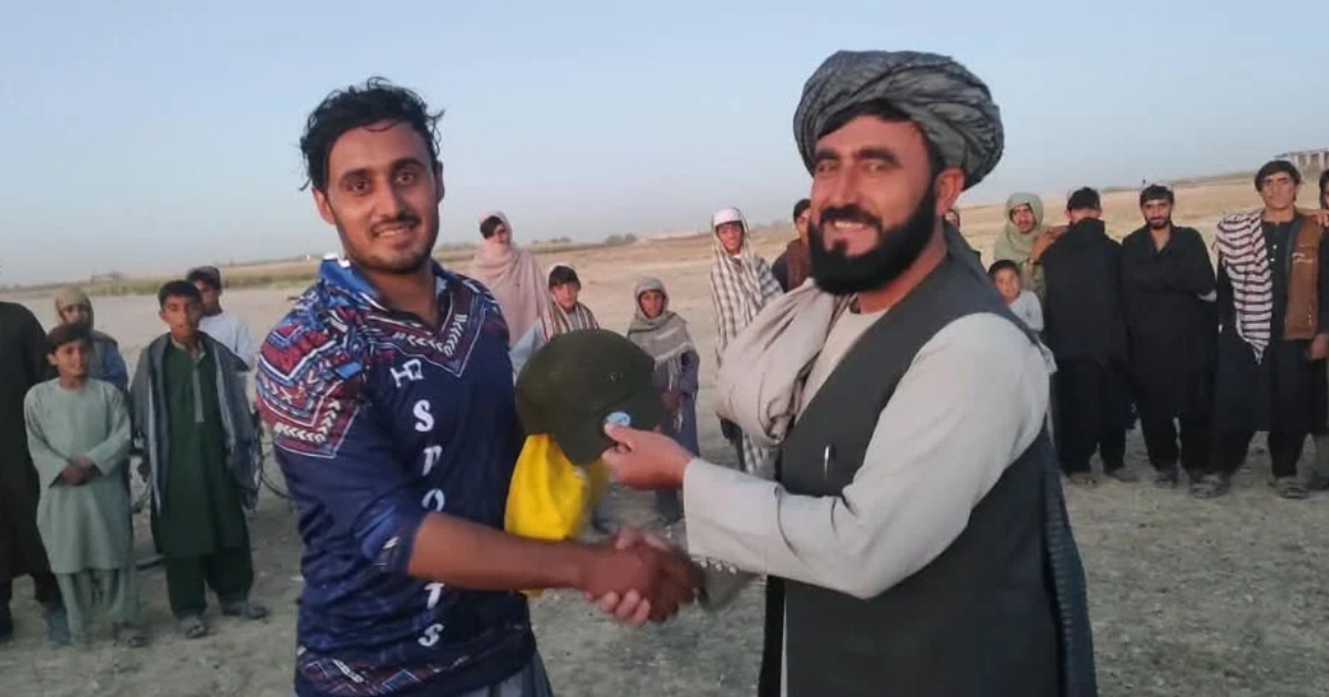 د کرکټ یو لوبغاړی په شین رنګه ورزشي لباس کې له یوه سړي سره د جایزې د سپارلو پر مهال روغبړ کوي او شاته یې نندارچیان ولاړ دي.