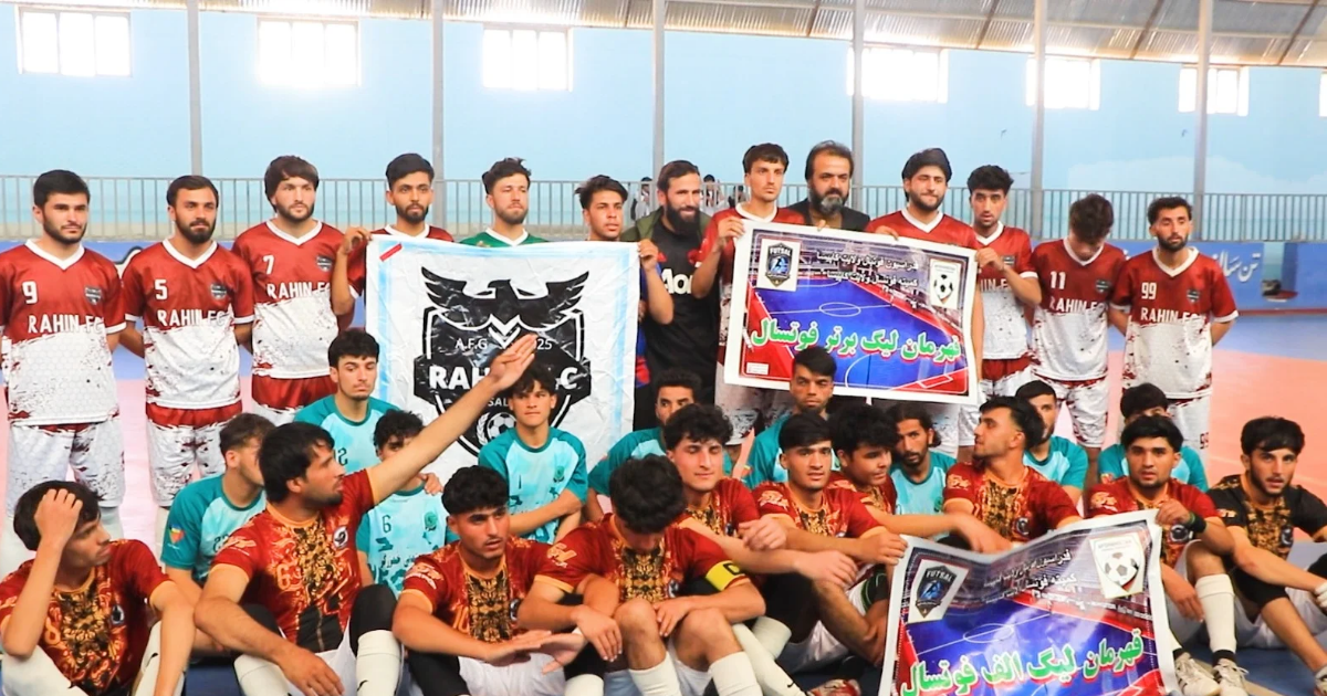 Players from several futsal teams pose together inside a sports hall in Kapisa, with Premier League and League A banners displayed.