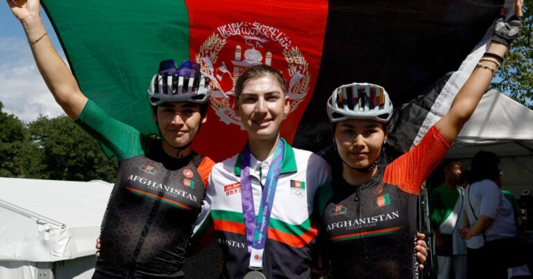 Fariba Hashimi wearing her Asian Games silver medal between two Afghan cyclists in front of the Afghan flag
