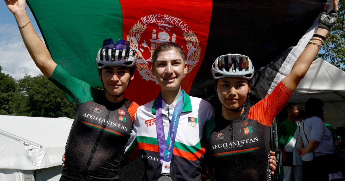 Fariba Hashimi wearing her Asian Games silver medal between two Afghan cyclists in front of the Afghan flag